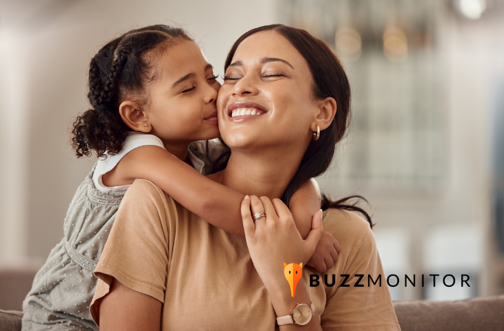 mãe e filha se abraçando no dia das mães. A criança tem um cabelo cacheado preso e está usando um vestido cinza. A adulta tem o caso liso longo, está sorrindo e usa uma blusa bege.